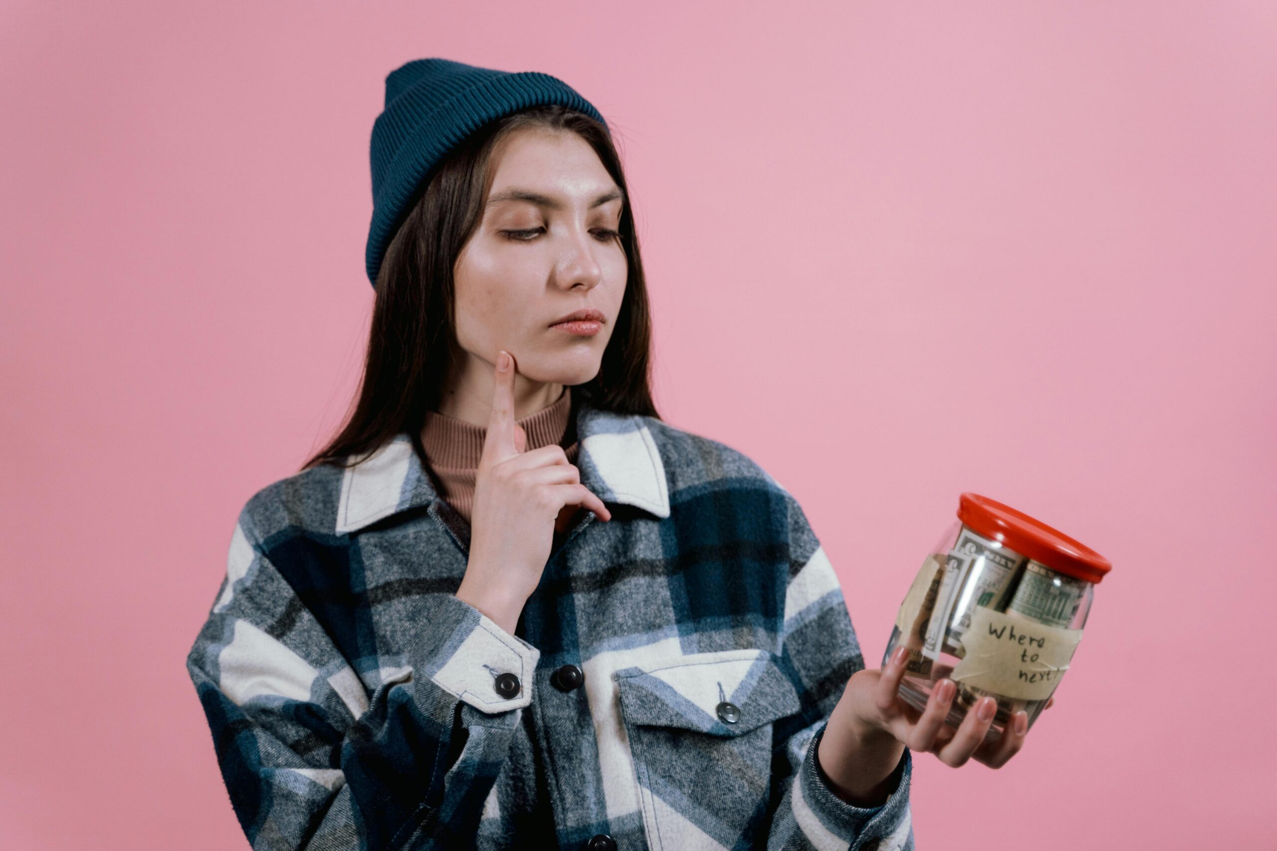 Pensive woman with a savings jar labeled 'where to next' on a pink background.